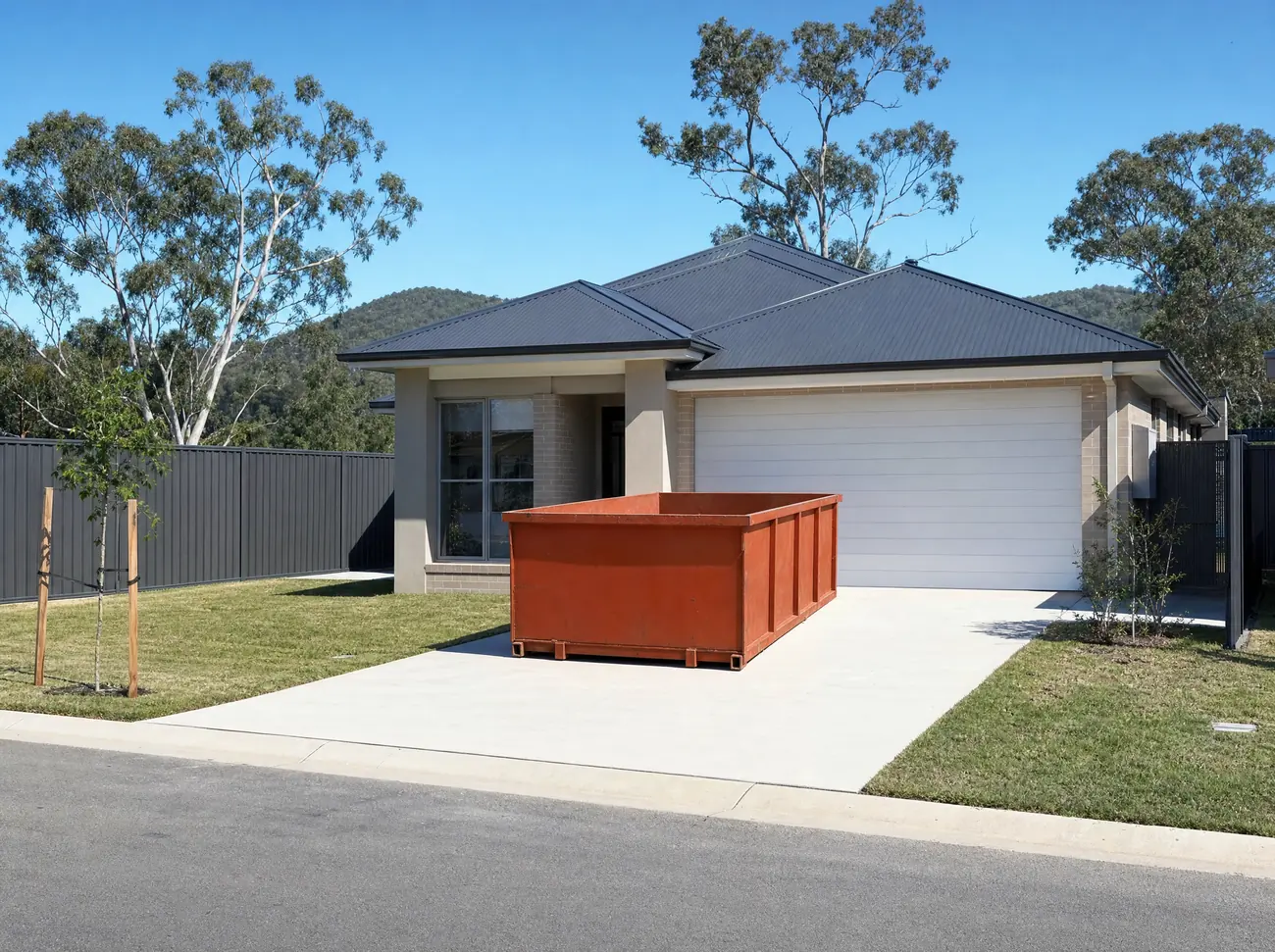 An empty orange skip bin sitting lengthways on the wide concrete driveway of a new rendered-brick home in a Maitland estate, fresh turf and a colorbond fence behind, young eucalypt street trees and low Hunter Valley hills beyond