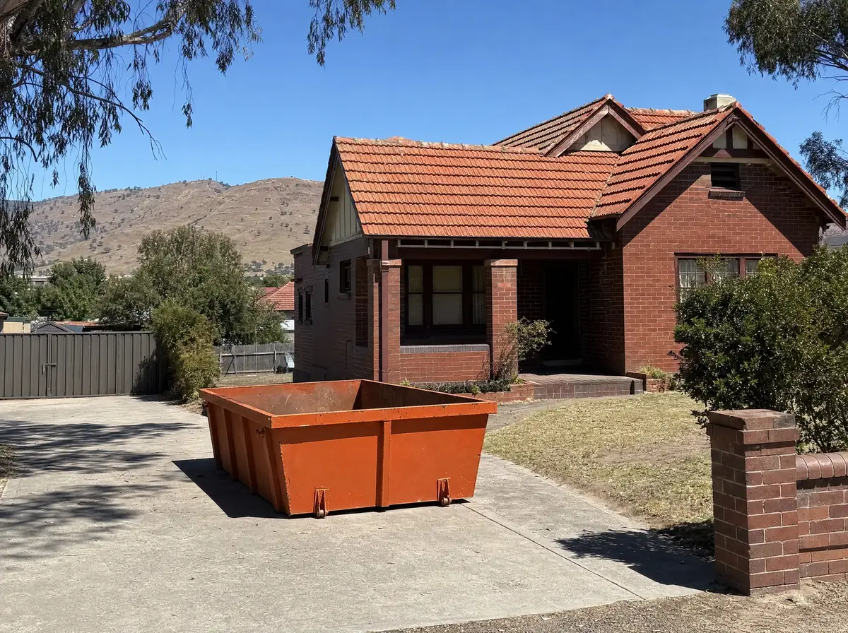 An empty orange skip bin sitting lengthways on the concrete driveway of an older red brick home in Muswellbrook, an old pepper tree in the front yard and dry brown hills rising close behind the town